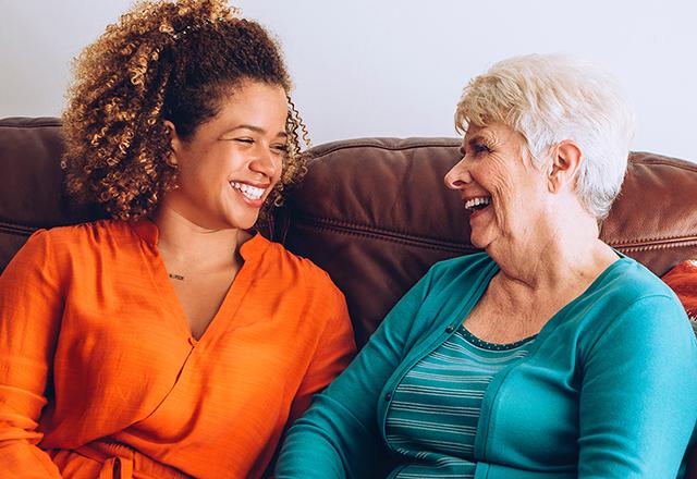 Older woman with short gray hair sitting on couch with younger woman and curly hair both laughing.